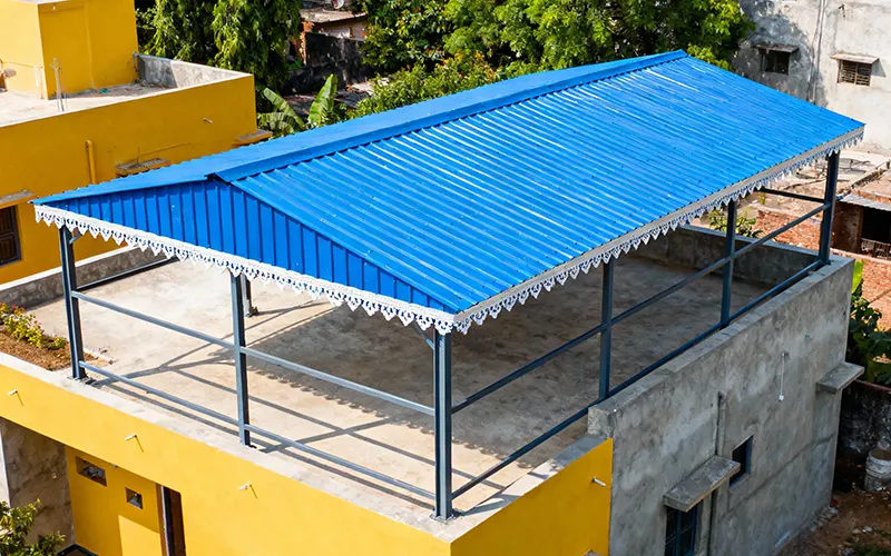 Indian residential house with a large blue metal rooftop shed supported by a steel frame, positioned on a modern concrete building. The shed features a single sloped blue corrugated metal roof with decorative edges, surrounded by greenery and trees. The scene is set in bright daylight within an urban neighborhood, highlighted by yellow walls and a mix of architectural styles.