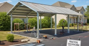 A large 20x30 shed with a gently sloping corrugated metal roof and robust steel columns, built in front of a residential house. The paved surface, green bushes, and visible construction tools illustrate ongoing shed installation work, while a clear blue sky with fluffy clouds provides a bright, practical setting focused on the construction process.